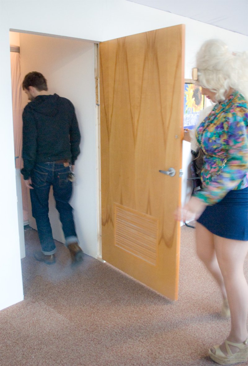 A person with blonde hair and a colorful shirt opens a wooden door for a person in a black hoodie and jeans.