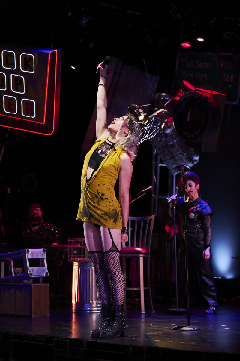 Blonde performer in yellow dress and fishnets sings into a microphone, arm raised, on a dark stage with neon signs and band members.
