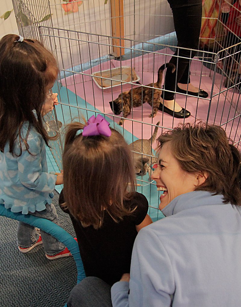 Two young girls and a woman in a light blue jacket laugh while looking at kittens in a cage with a pink floor.