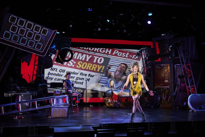 Drag queen in yellow dress, fishnets, and boots on stage, with large screens displaying newspaper headlines and a man in the background.