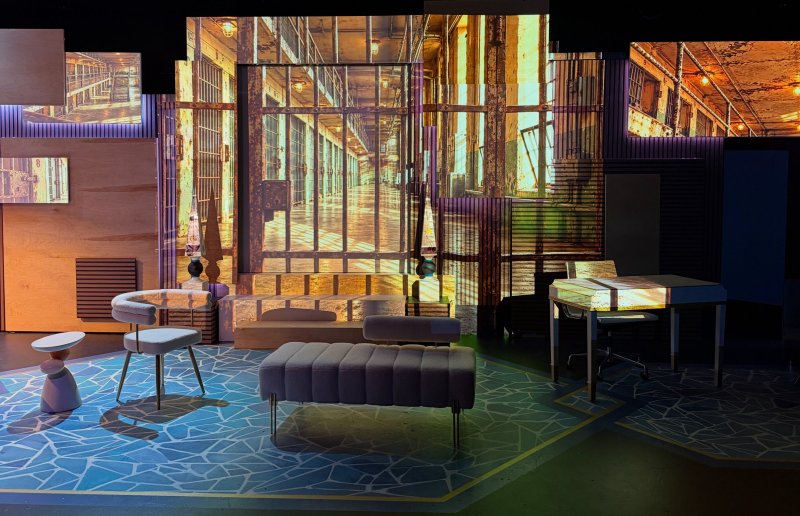 Multimedia installation with projected prison hallway on screens, furniture, and blue geometric floor patterns.