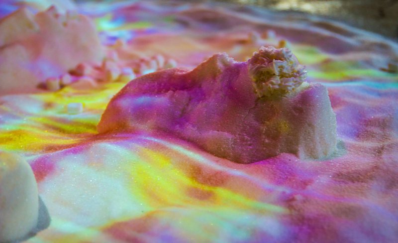Close-up of a sand-like landscape with pink, purple, yellow, and blue light projected onto it, creating a shimmering effect.
