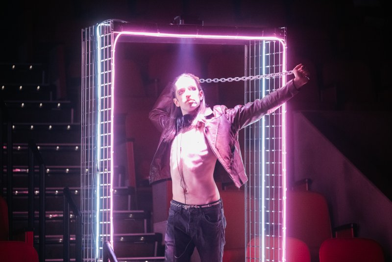 Performer in open leather jacket and jeans, chained to a glowing pink and white neon frame on a dark stage.