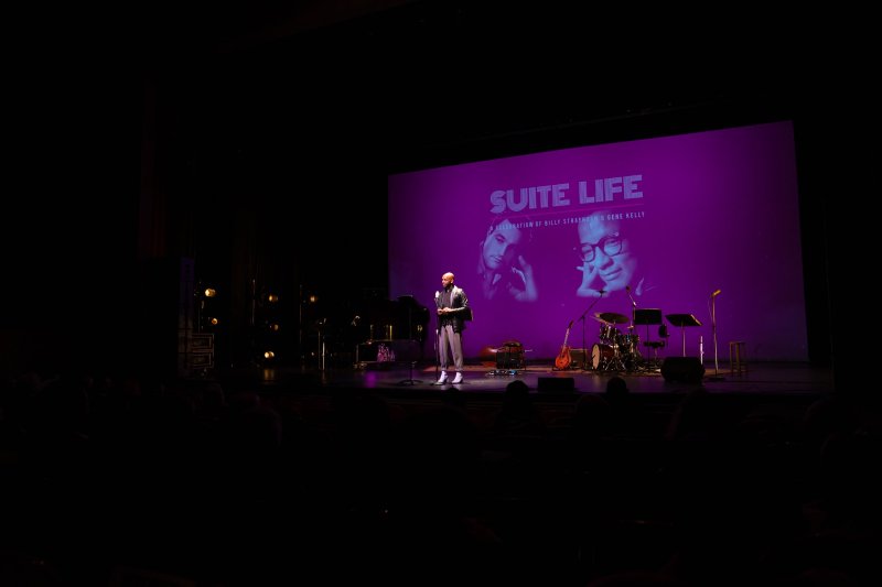 A person in a red hat and black clothing stands on a stage, speaking into a microphone, with a purple "Suite Life" screen behind them.