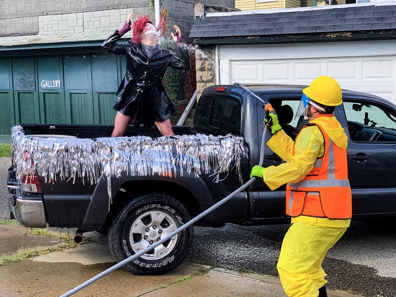 Person in yellow hazmat suit sprays water on a performer in a black coat and red fishnets standing in a truck bed.