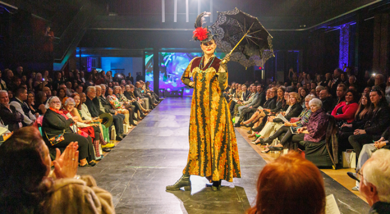 A model in a long, patterned dress, elaborate hat, and holding a lace umbrella walks a runway before a seated audience.