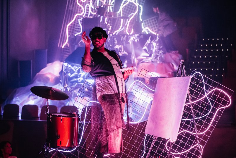 Man in hat and sunglasses on stage with drum, cymbal, easel, and neon grid lights, bathed in purple and pink light.