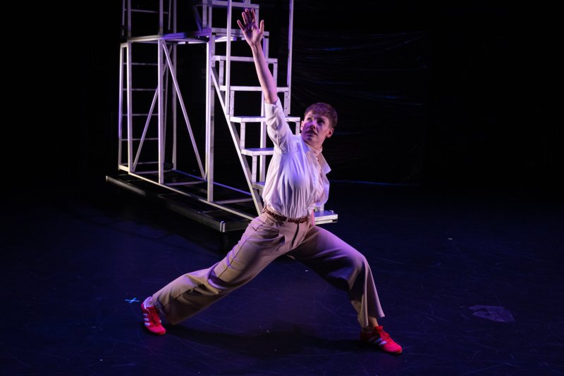 Dancer in white shirt, tan pants, and red shoes in a lunge, right arm raised, on a dark stage with white scaffolding.
