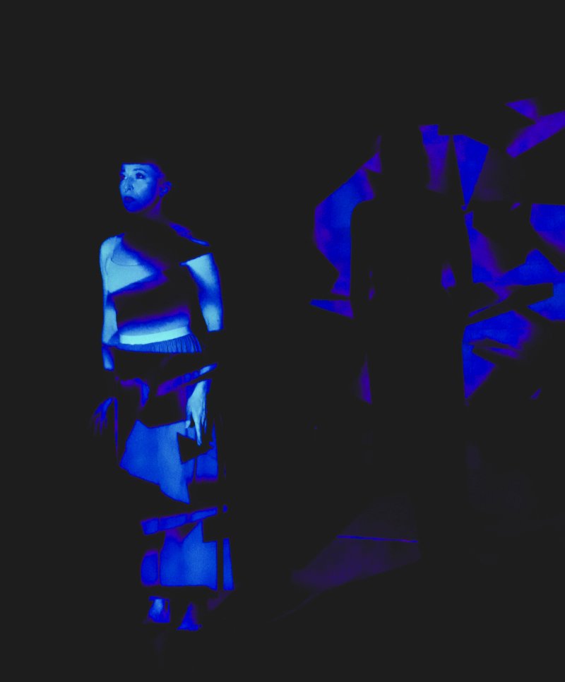 A dancer in blue light, with geometric patterns projected onto their body, stands against a dark background.