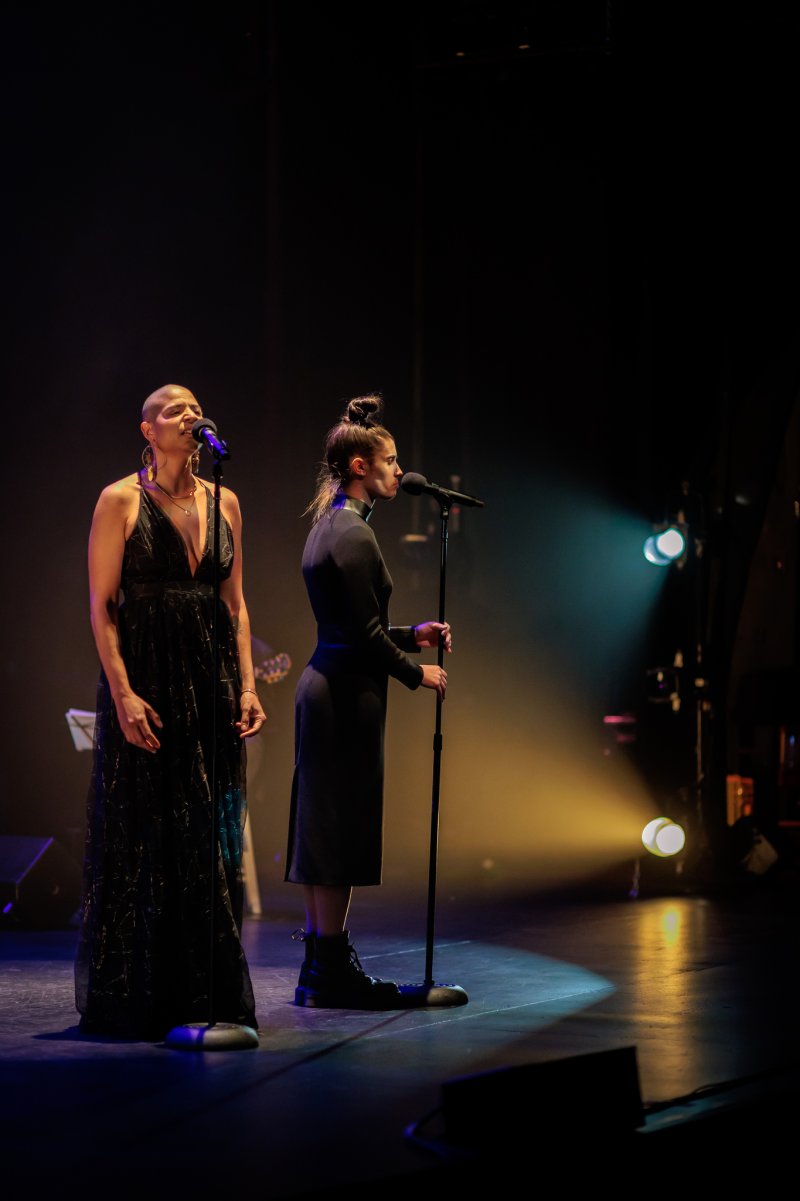 Two women singing on a dark stage, illuminated by spotlights. One wears a long, dark gown, the other a dark dress and boots.