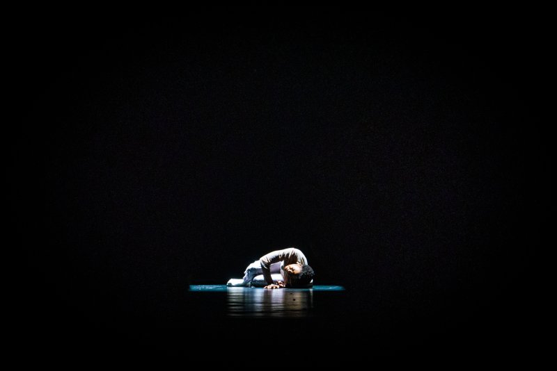 Dancer in white, curled on a reflective surface under a spotlight, against a dark background.