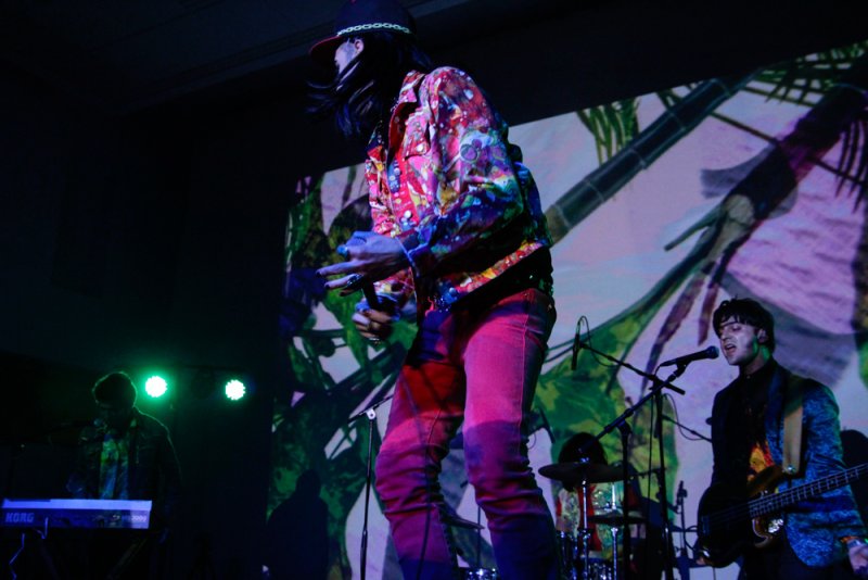 Musician in a colorful jacket and red pants on stage, performing with a band against a projected abstract background.