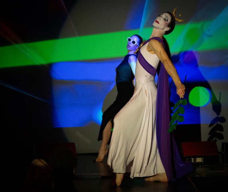 Two performers on stage. One in white and purple with a crown, holding a green branch. The other in black with white sunglasses.