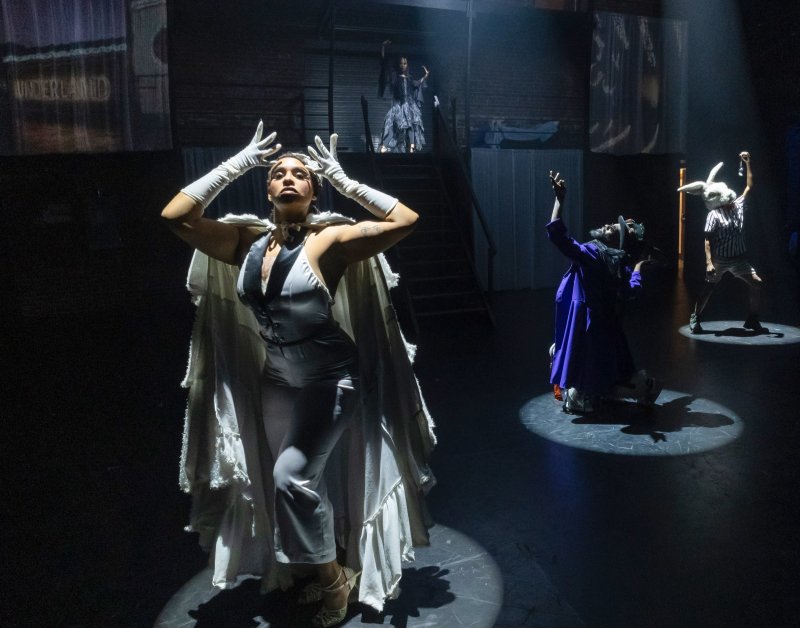 Staged performance with figures in theatrical lighting. A central figure in white gloves and cape stands out, with others including one in a rabbit mask, and a backdrop with the word "Underland.