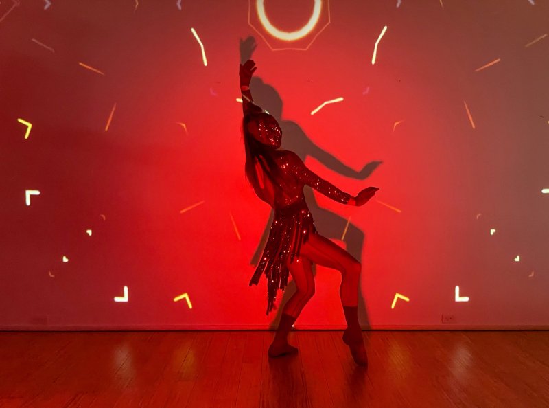 Performer in a shiny red suit striking a pose against a red wall with projected white geometric patterns and a large circular halo.