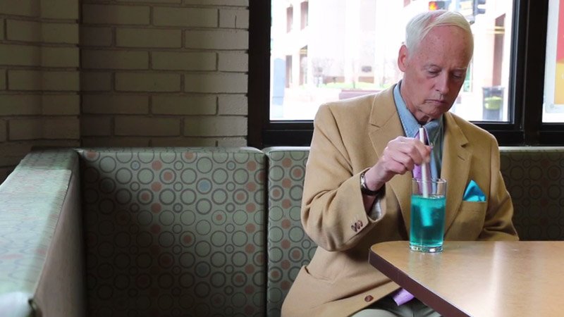 A man in a tan suit stirs a blue drink with a purple object in a diner booth.