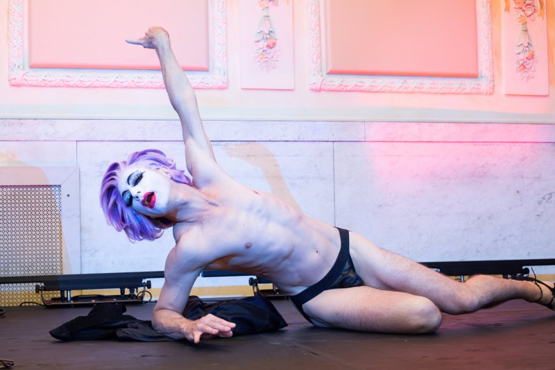 Performer with purple hair and white face makeup reclines on a stage, wearing a black jockstrap. Arm raised, pointing. Pink and blue lighting, ornate wall details in background.