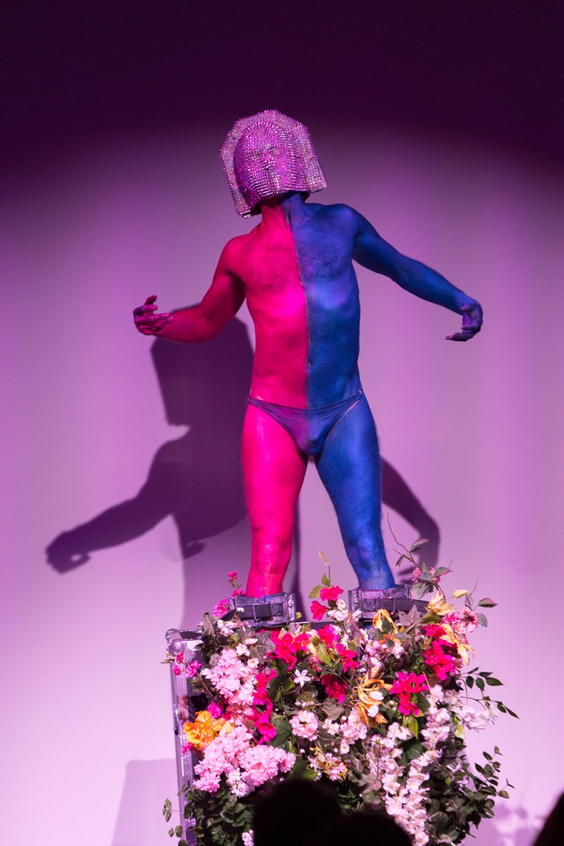 Performer Darrell Thorne in a glittery helmet, half pink and half blue body paint, standing in a flower-filled box.