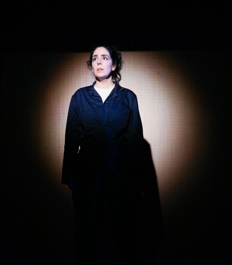 Woman in dark blue jumpsuit, lit by a spotlight, looking up with a worried expression against a dark background.