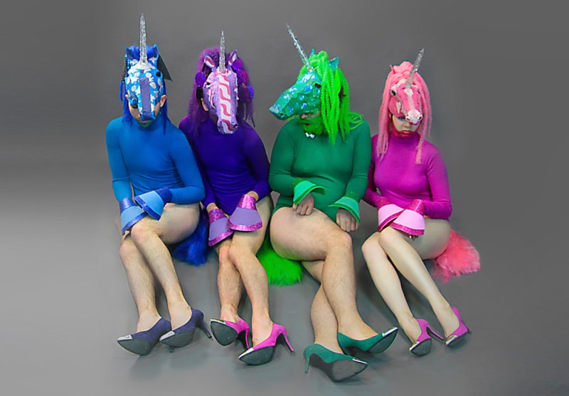 Four people in colorful unicorn masks, bodysuits, high heels, and furry tails sit against a gray background.