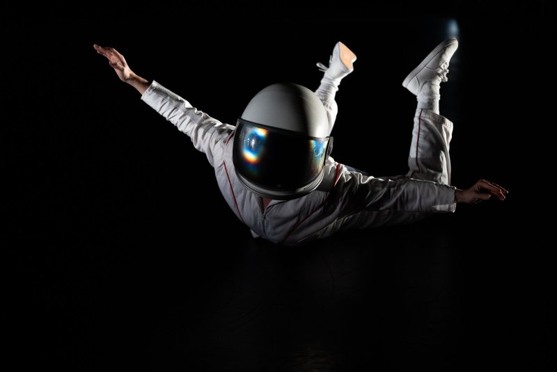 Astronaut in white suit, helmet reflecting light, floating against a dark background with arms outstretched and legs bent.