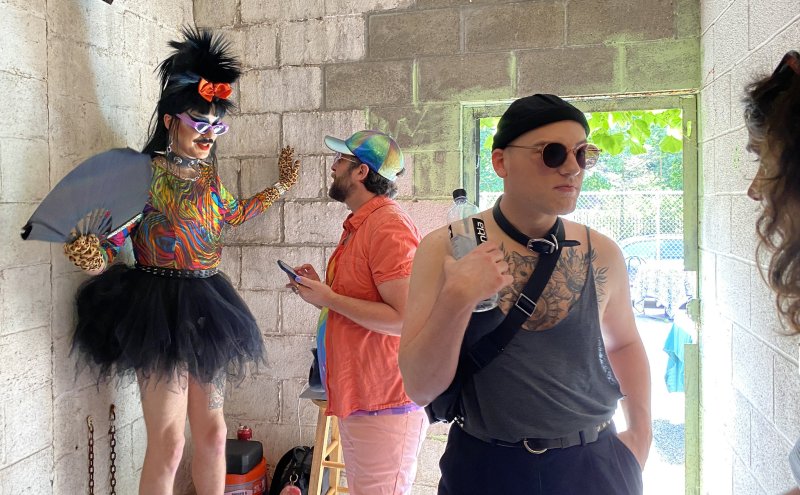 Drag queen in colorful outfit, black tutu, and spiky hair poses next to two men in a concrete block room.