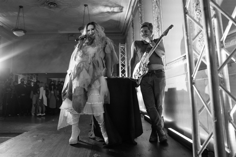 Black and white photo of a drag queen singing into a microphone next to a guitarist on a stage.