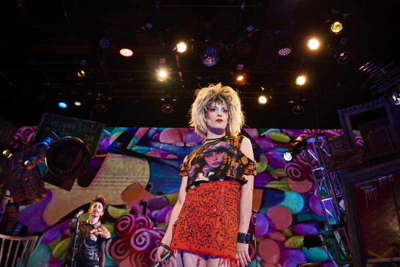 A blonde performer in a cropped t-shirt and red skirt stands on stage under bright lights, another performer behind them.