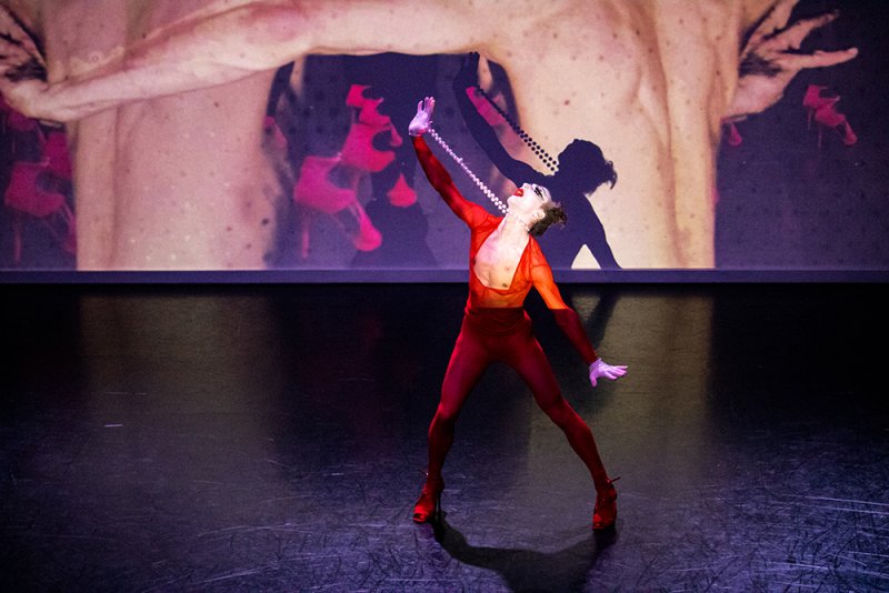 Performance art piece featuring a dancer in a red bodysuit and heels, holding a beaded necklace. A large projection behind shows a torso and floating red shapes. The stage is dark.