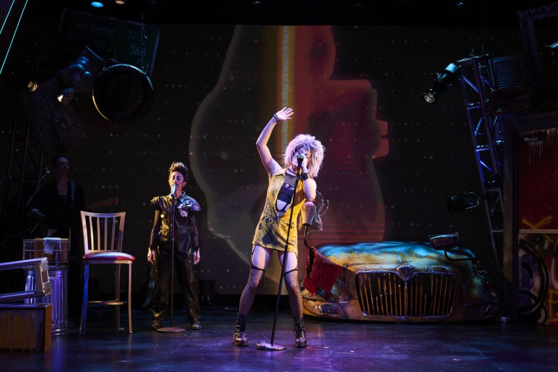 Scott N Andrew's "Hedwig and the Angry Inch" (2026): A performer with blonde hair and a yellow dress sings into a mic on a stage with a band and car prop.
