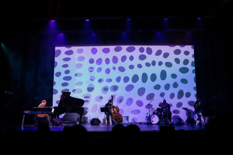 Jazz band on stage with piano, bass, and drums, performing in front of a large screen displaying purple and white abstract shapes.