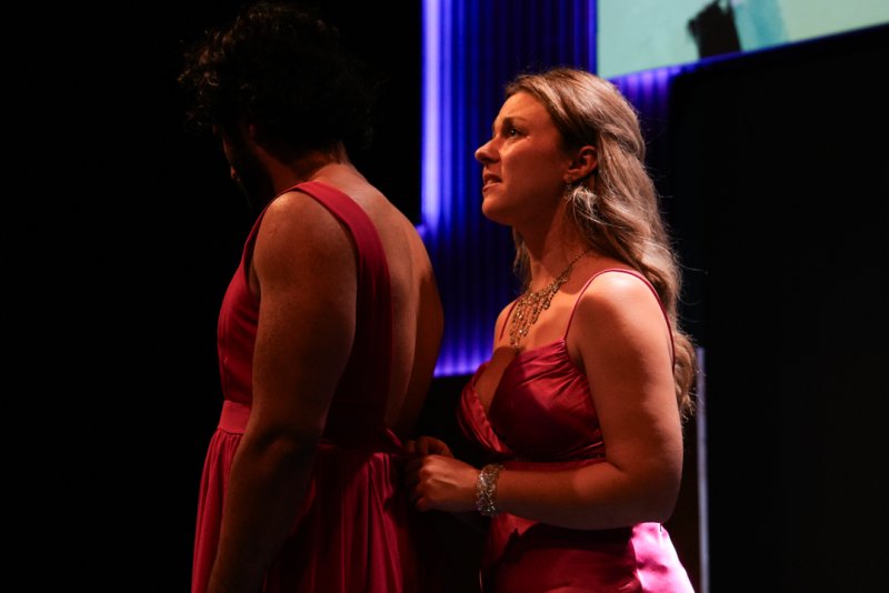 Two women in pink dresses on a dark stage; one faces away, the other looks up, adjusting a strap.