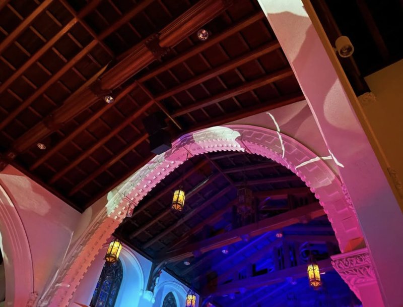 Projected light on a white archway in a church, with a dark wooden ceiling, hanging lanterns, and blue light illuminating the background.