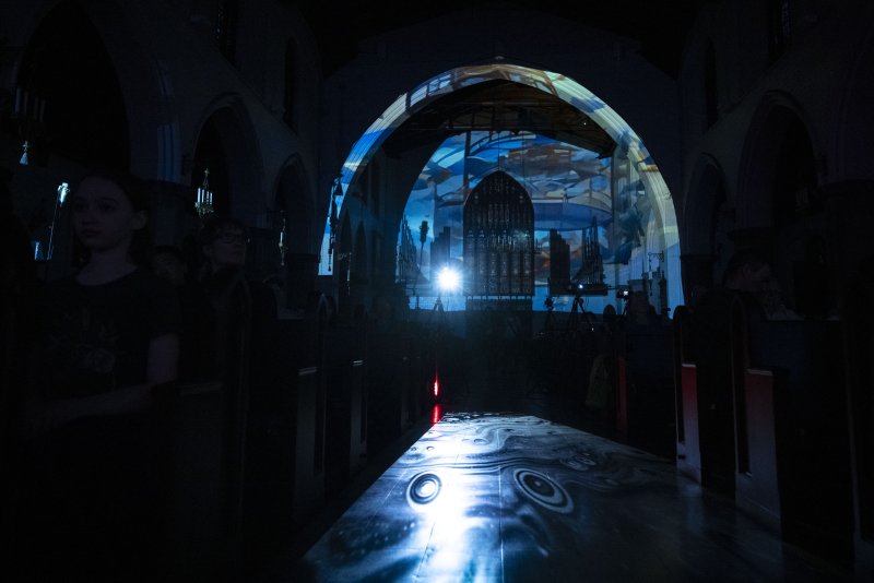 Dark church interior with a bright blue and white light projection on the altar and floor, showing abstract patterns and cityscapes.
