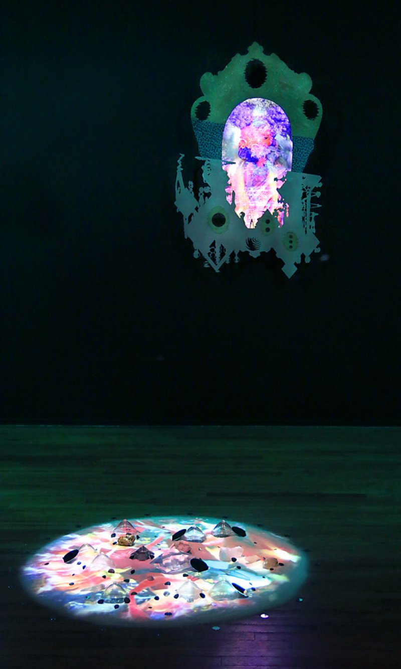 Digital projection of a glowing, purple and pink face on an ornate, white cutout, above a spotlighted pile of crystals.