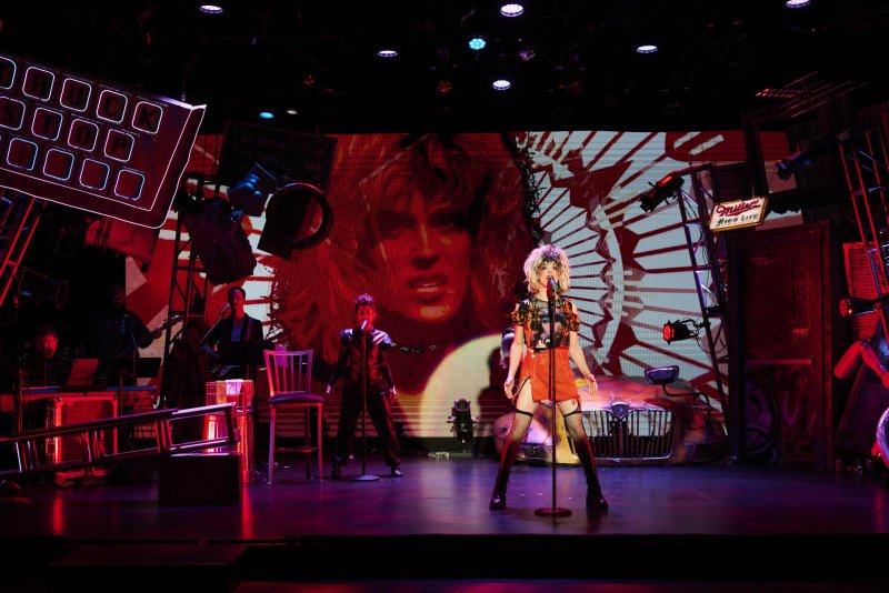 A blonde singer in an orange dress and fishnets stands center stage, backed by a large screen displaying a woman's face.