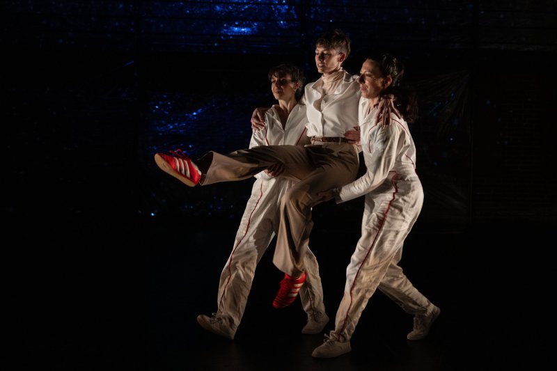 Three dancers in white and tan outfits, one held aloft, against a dark, textured blue background.