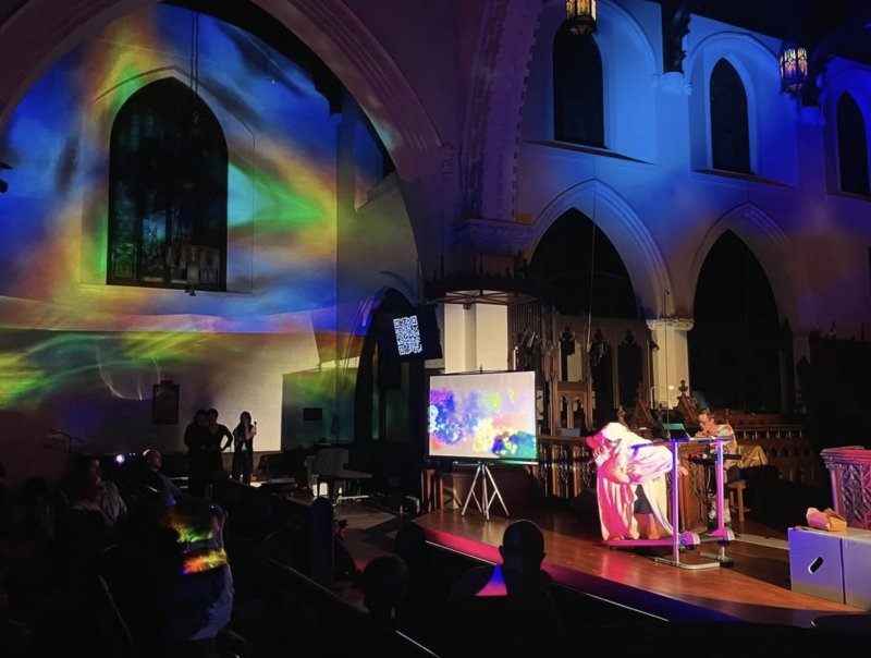Performance in a church with colorful light projections, a screen displaying abstract art, and a performer in a flowing white costume.