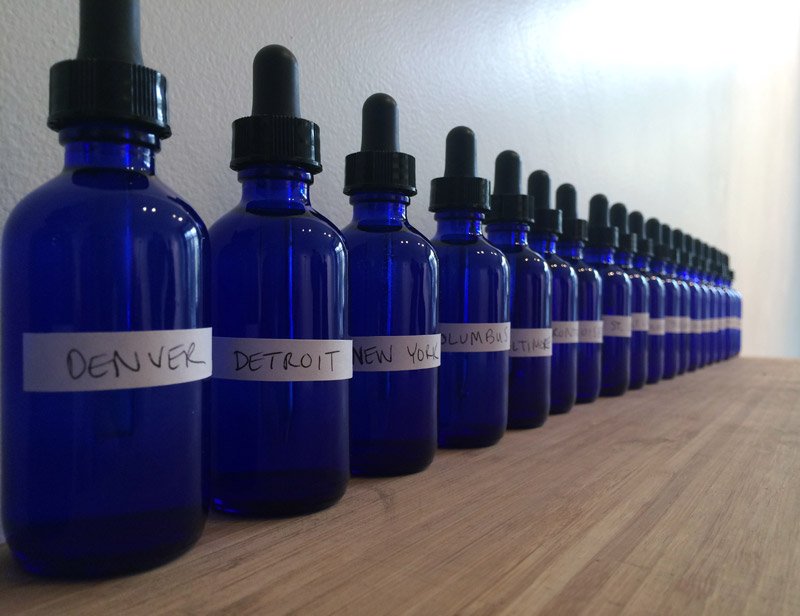 Row of blue dropper bottles with white labels, each bearing a city name, on a wooden surface against a white wall.