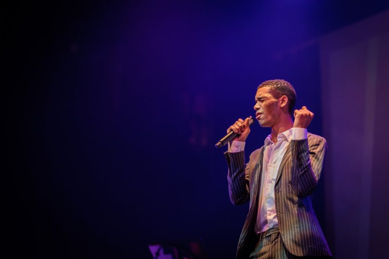 Man in pinstripe suit singing into a microphone on a dark stage, illuminated by purple light from the left.