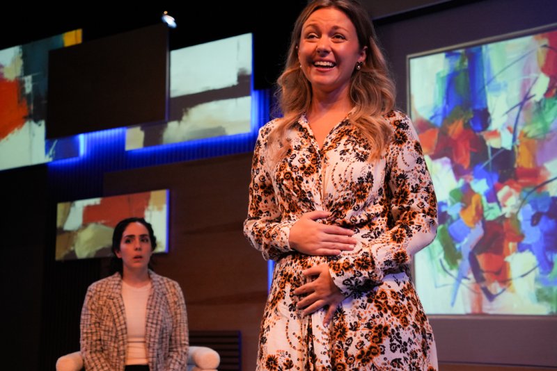 A woman in a floral dress laughs on stage, another woman sits behind her, abstract art on screens in the background.