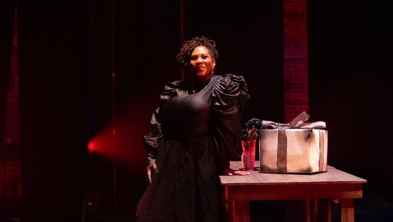 Portrait of a Black woman in a black dress, standing by a table with a gift and flowers, lit by red light.