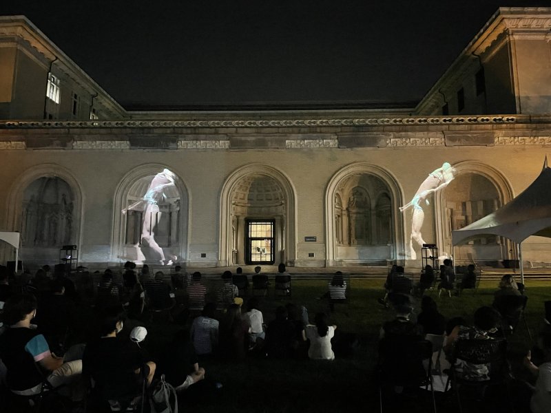 Two large, ghostly, female figures projected onto an arched building facade at night, with an audience seated on grass below.