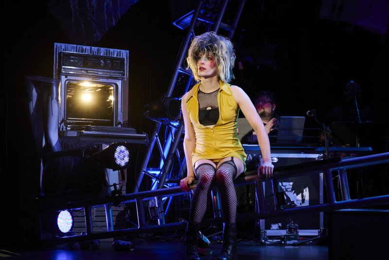A person in a yellow dress, fishnets, and a blonde wig sits on a ladder on a dark stage with bright lights.