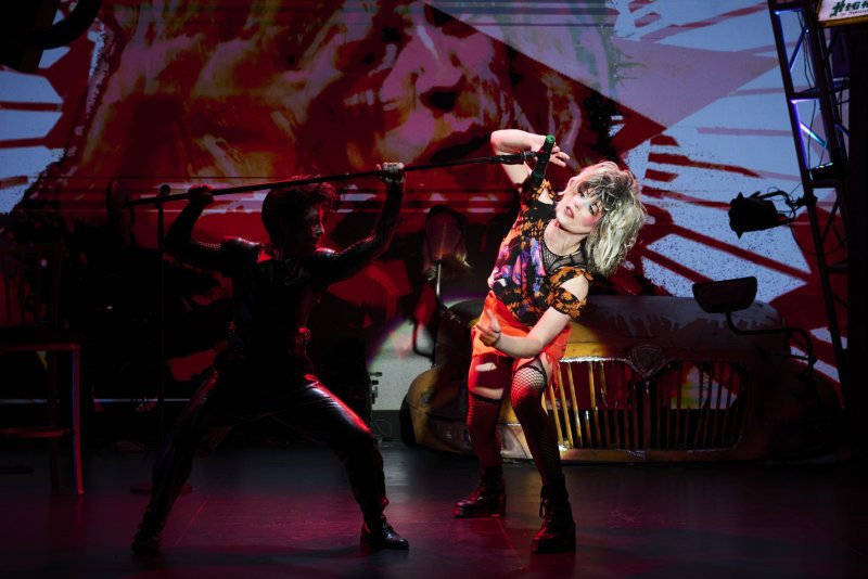 Two performers on stage: one in black leather holding a staff, the other in a colorful top and fishnets singing into a microphone.