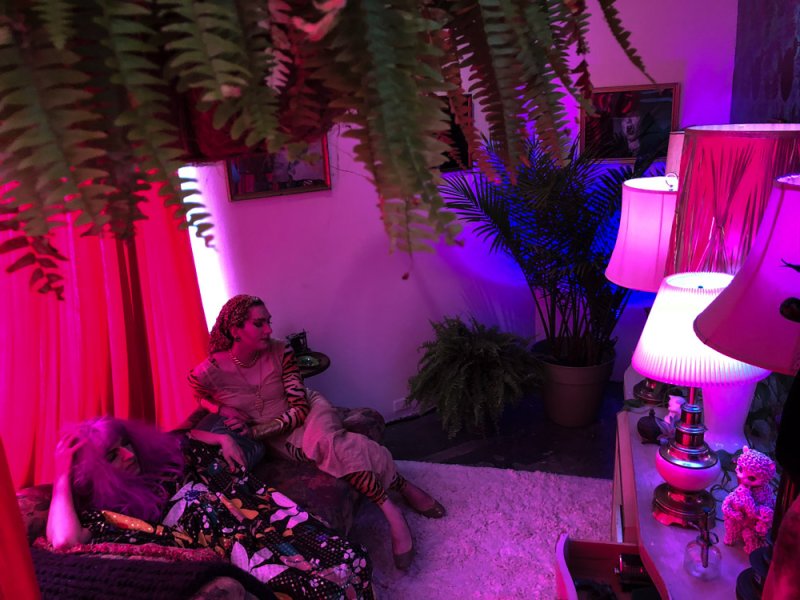 Two figures in a room bathed in pink and purple light, one reclining on a patterned couch, the other seated beside.