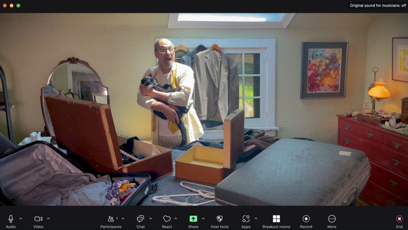 Man in yellow apron holds black cat, mouth open, surrounded by open suitcases and clothes in a sunlit room.