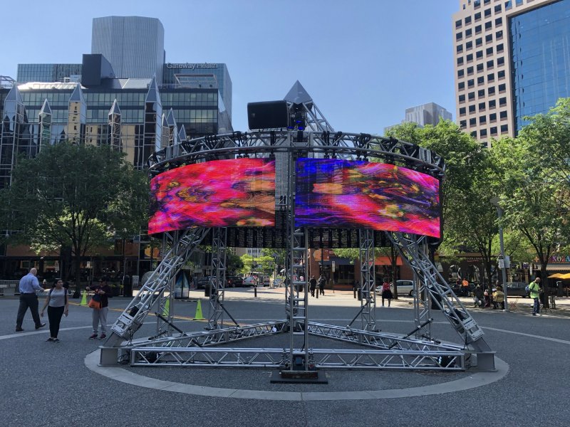 Outdoor art installation of two curved LED screens displaying abstract red and blue patterns, supported by a steel frame.