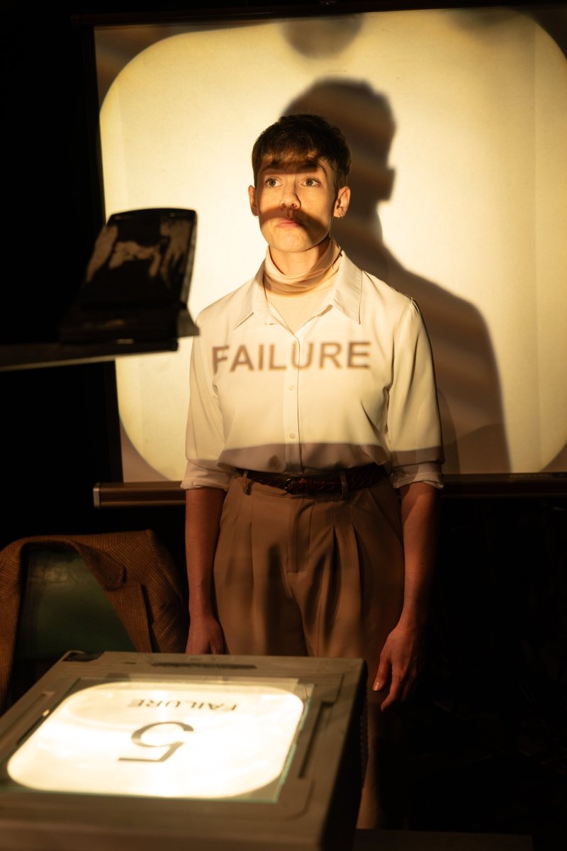 Man in white shirt and tan pants with "FAILURE" projected on his chest, standing before a projector screen.