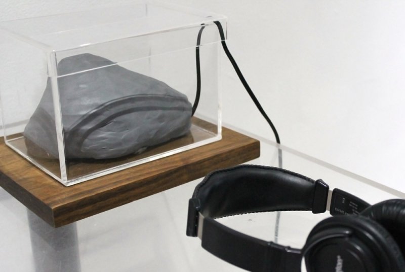 Gray felt rock in clear box on wood base, connected by black cord to black headphones on clear table.
